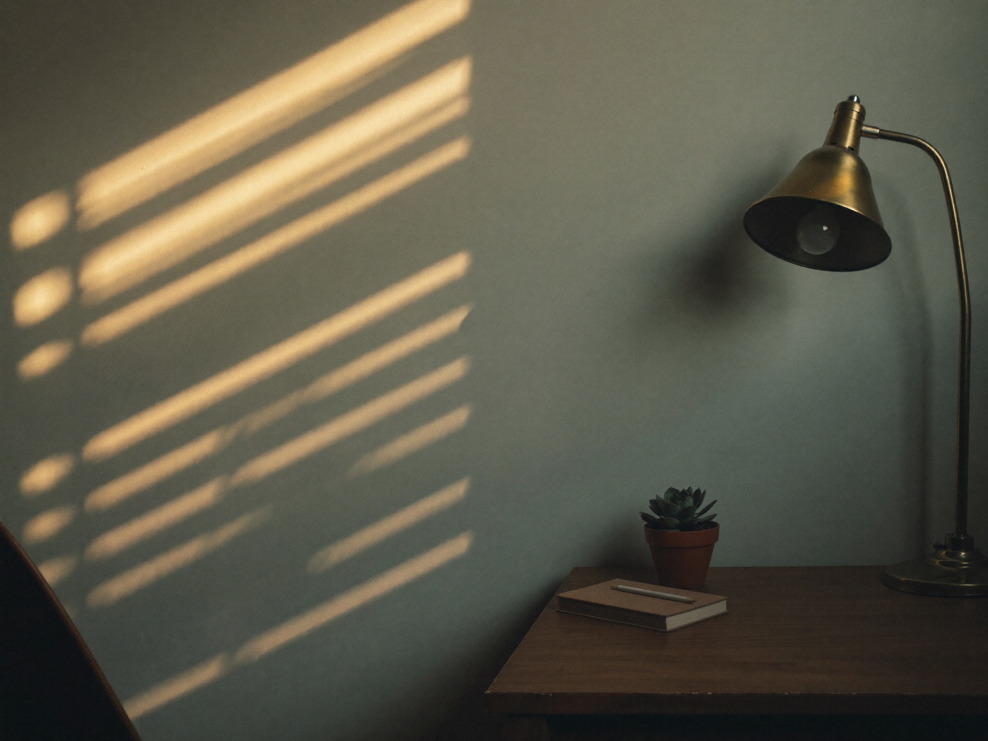 A minimal study corner at sunrise: a closed book, a pencil and slatted window light on a bare wall